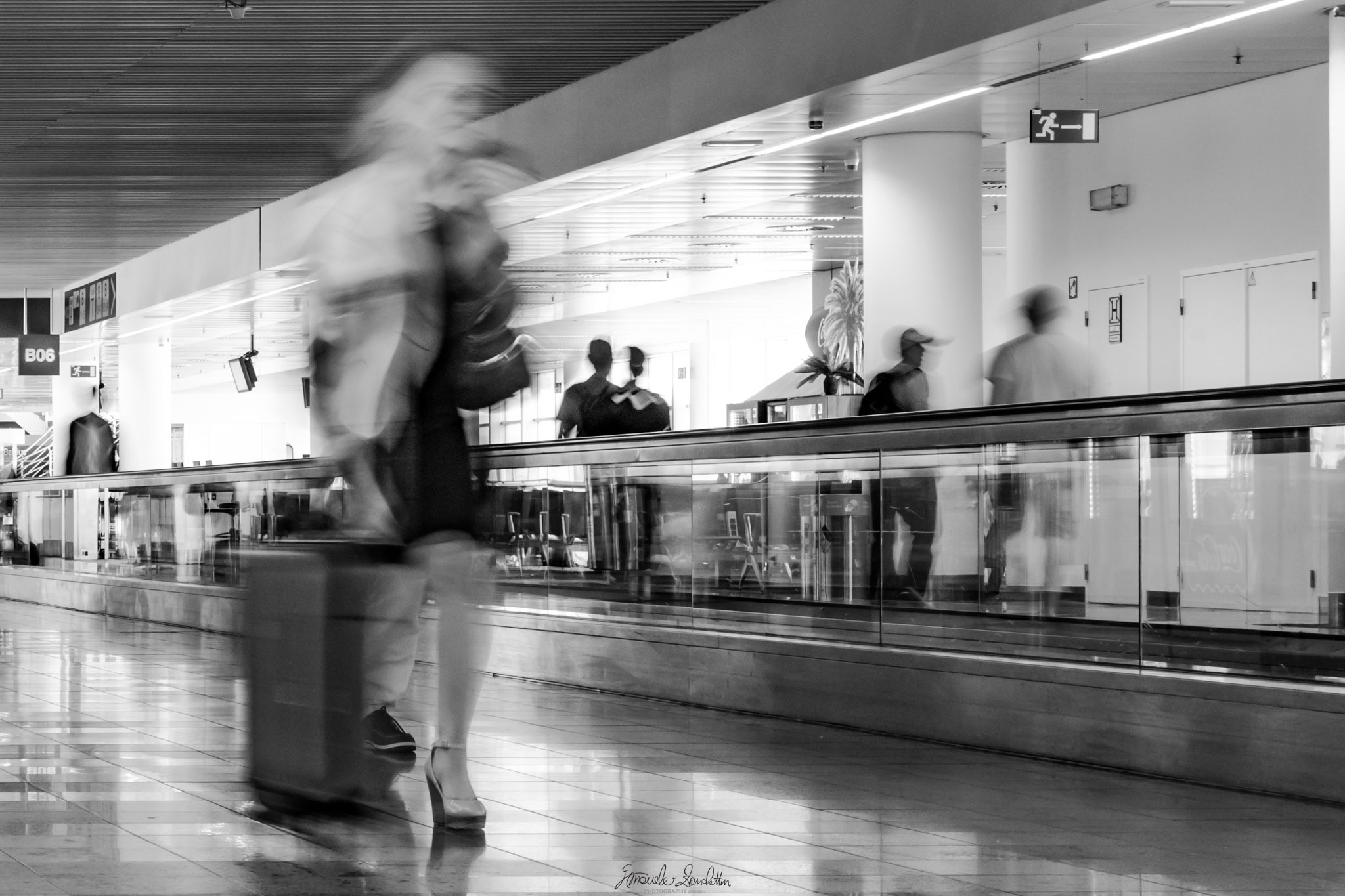 Black-and-white airport scene in which travellers blur during a slow exposure while signs, columns and the moving walkway remain defined