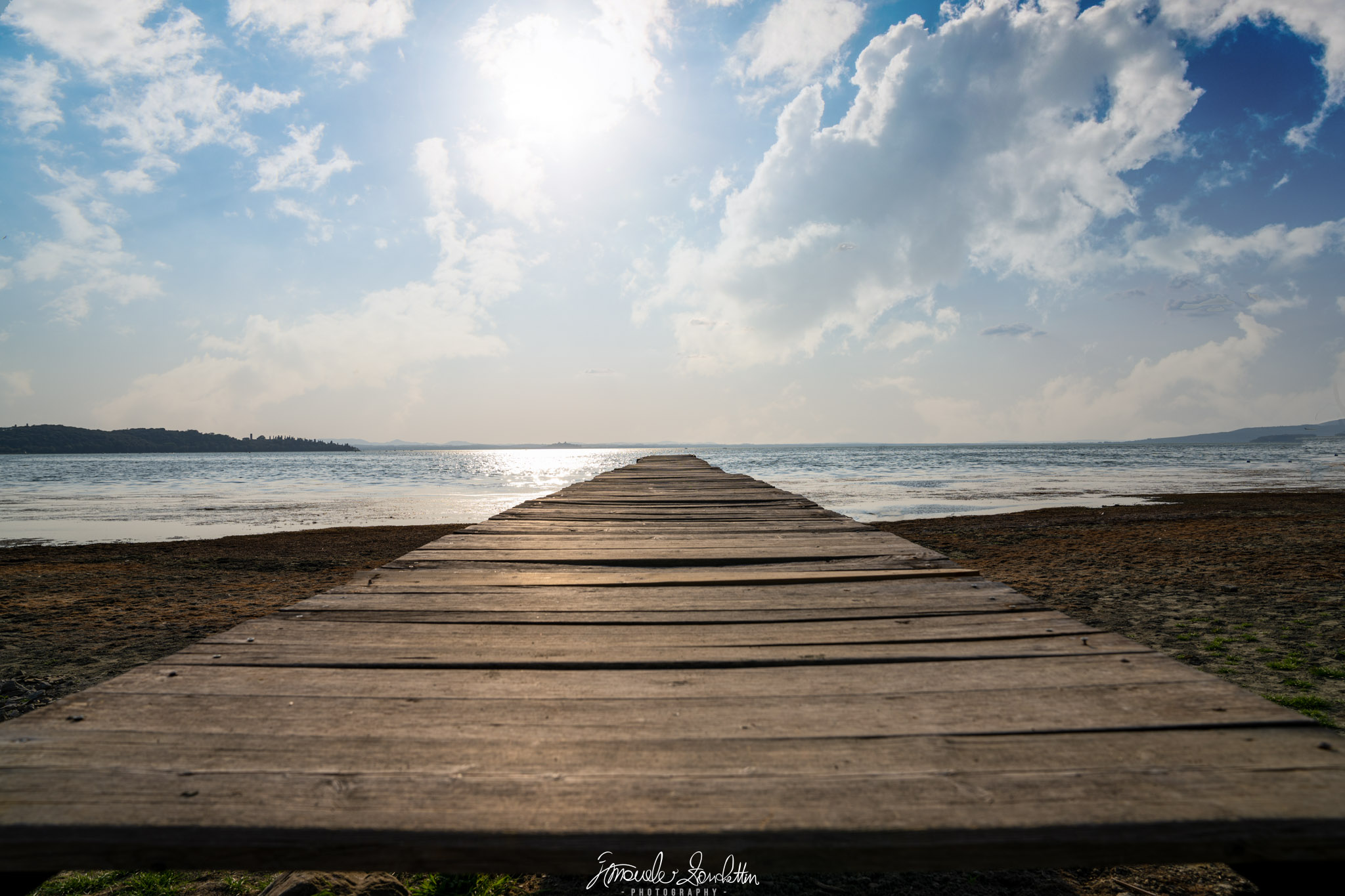 A broad wooden jetty narrows towards open water beneath a blue, cloud-filled sky. Bright sunlight and its reflection sit to the left of the jetty, with distant land on the horizon.