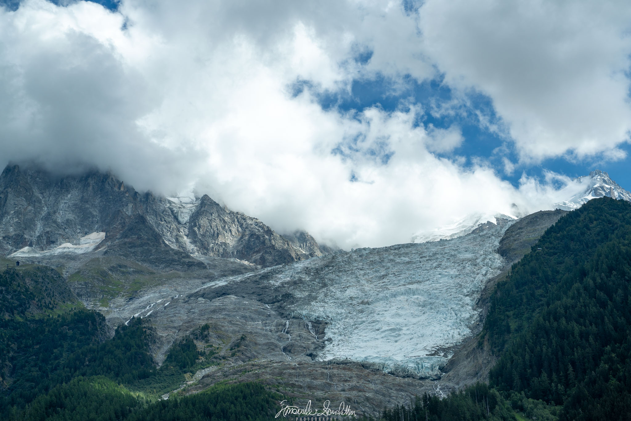 Un ghiacciaio chiaro curva tra versanti rocciosi e alberi scuri, sotto grandi nuvole e una porzione di cielo azzurro.