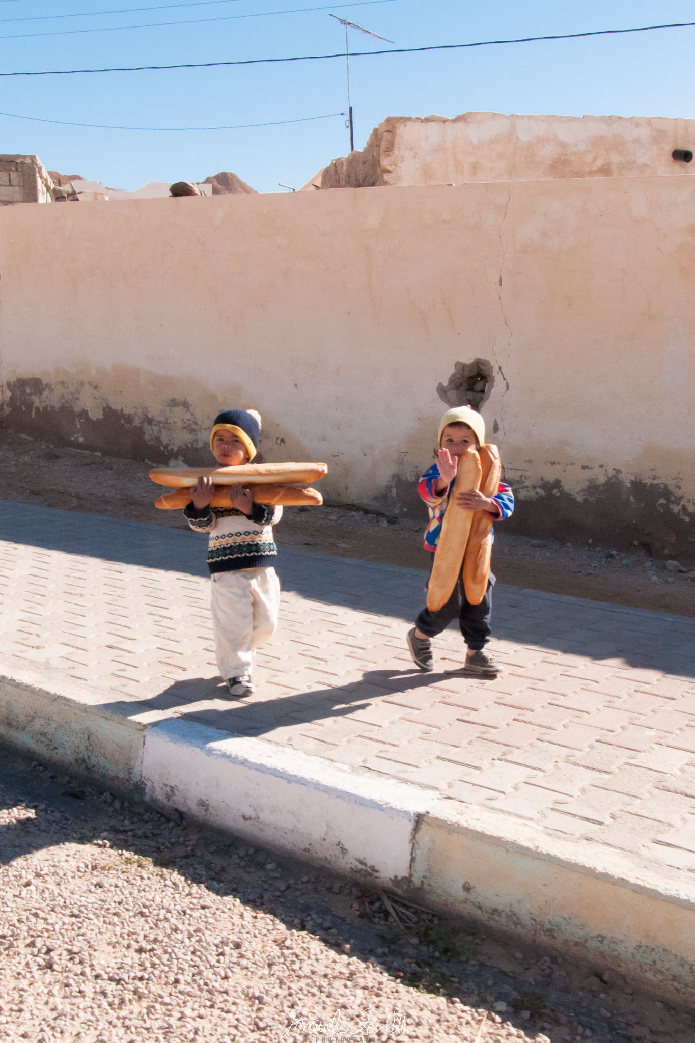 Two children carrying long loaves of bread stand beside a weathered wall. One raises a hand; strong shadows fall across the ground.