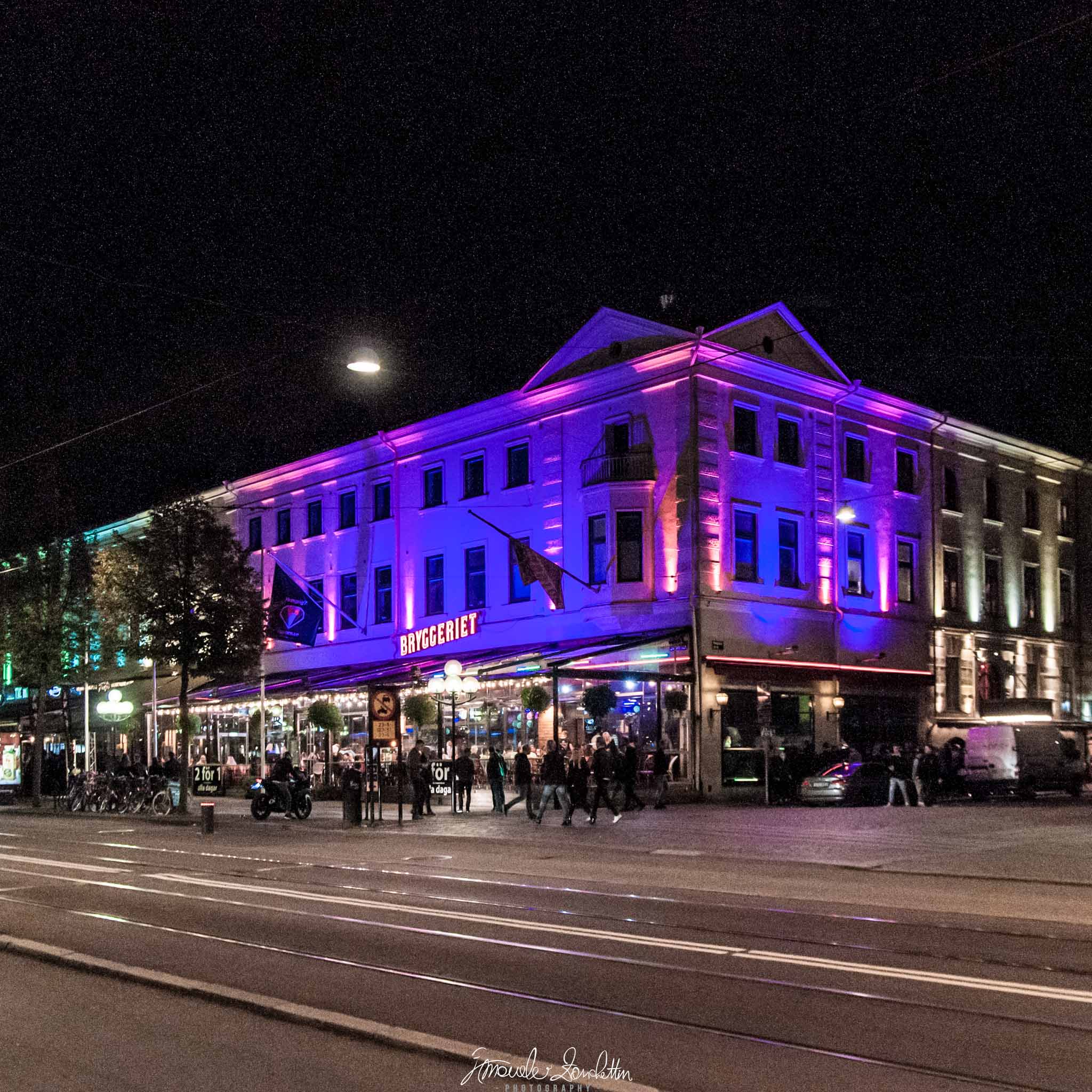 Un edificio d’angolo illuminato di viola e magenta di notte, con persone vicino all’ingresso, un lampione sospeso, binari e un veicolo sul bordo destro.
