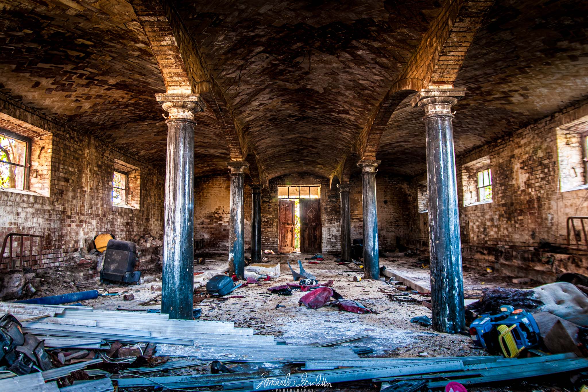 Un interno segnato dal tempo con volte di mattoni ripetute, colonne blu, finestre laterali luminose e materiale sparso sul pavimento, verso una porta centrale.