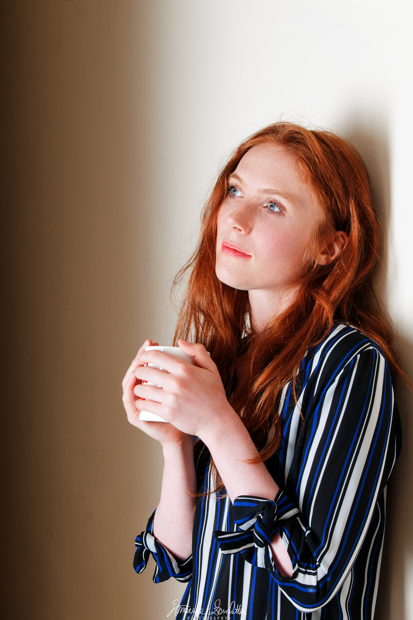 Una donna dai capelli rossi tiene una tazza bianca con entrambe le mani e guarda in alto a sinistra, accanto a una parete chiara, indossando una camicia scura a righe.