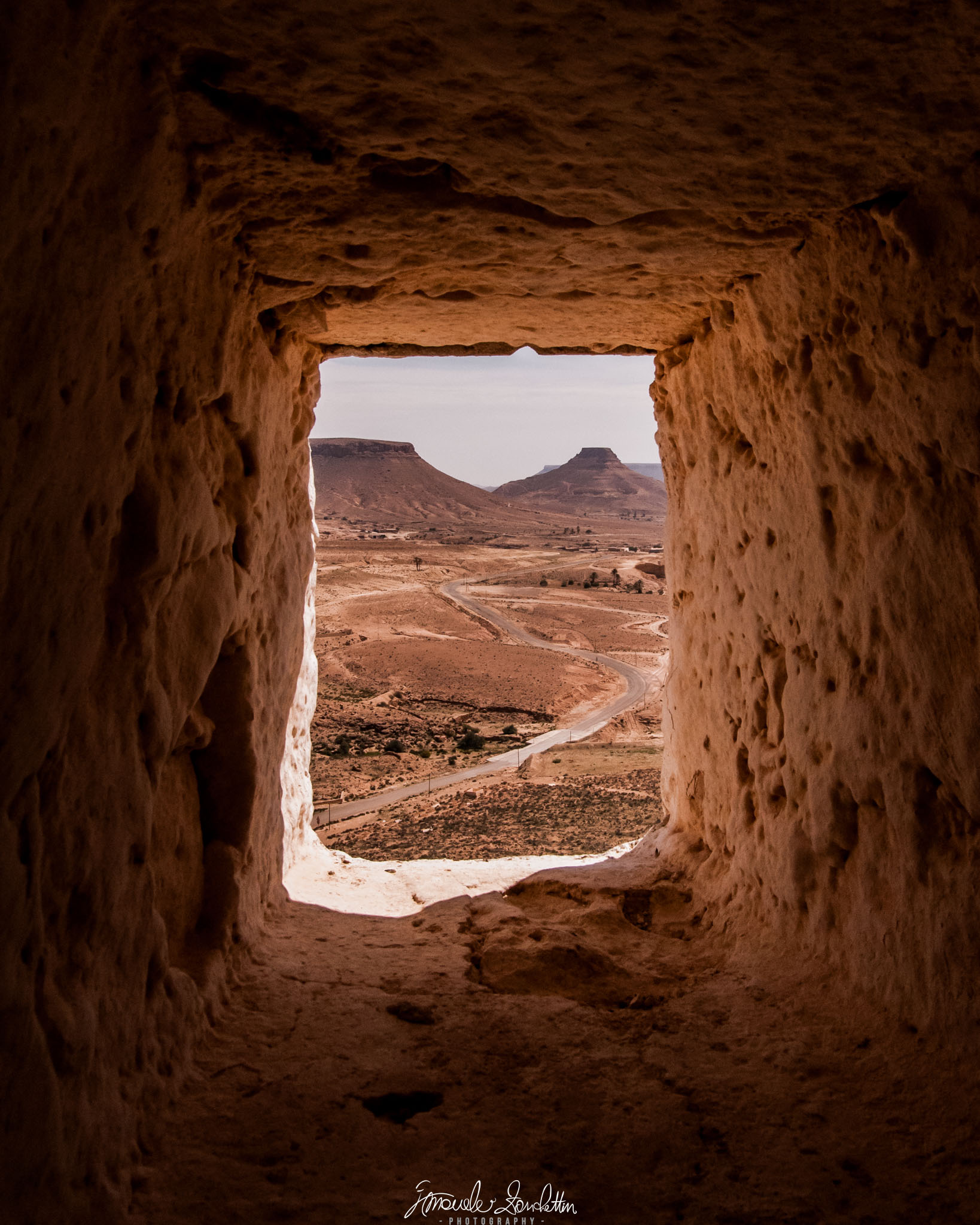 Un’apertura profonda e irregolare in una parete color terra incornicia una strada sinuosa su terreno arido verso rilievi dalla cima piatta.