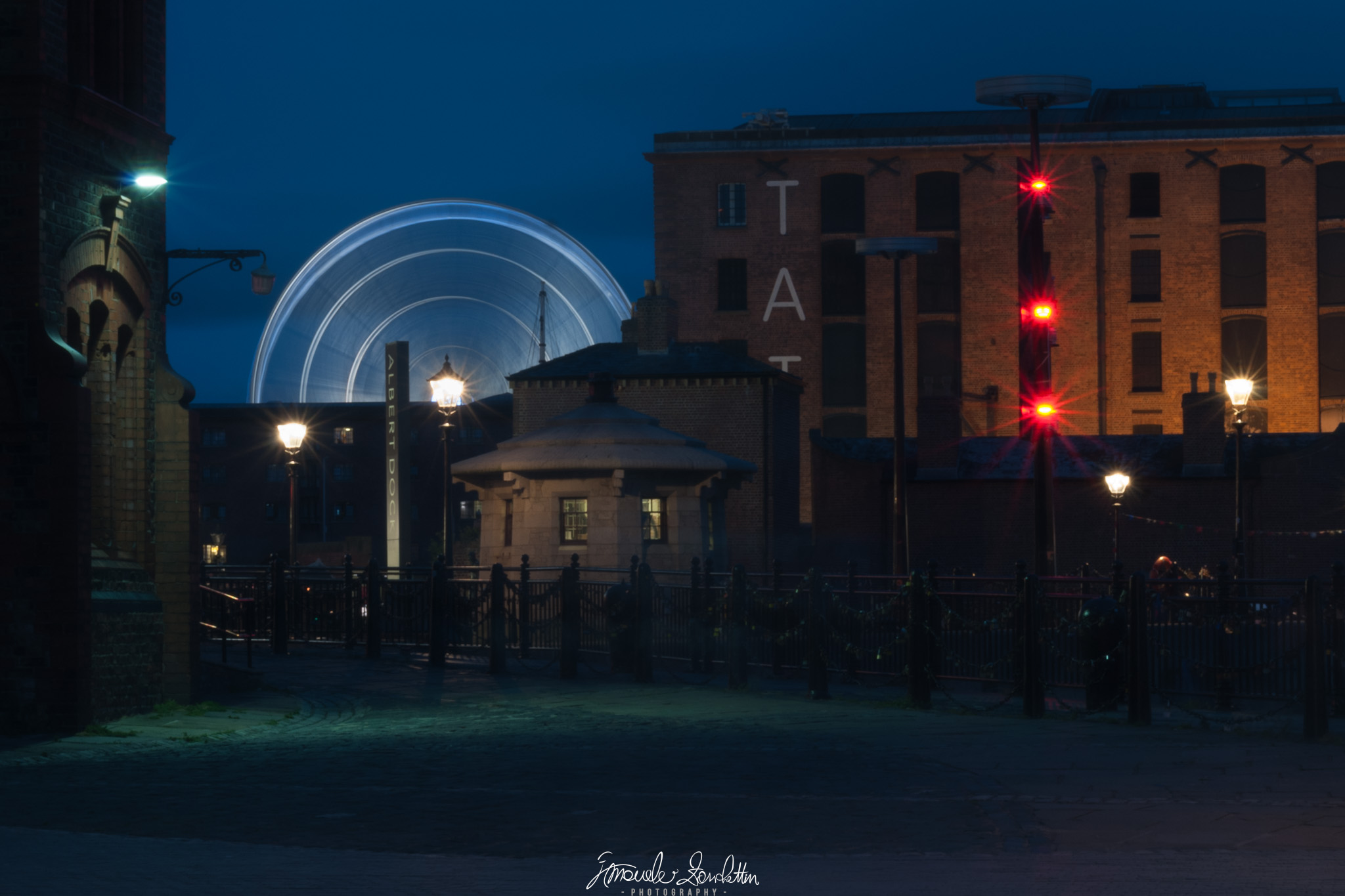 Scie luminose circolari di una grande ruota emergono dietro edifici scuri, con lampioni e luci rosse contro un cielo blu intenso.