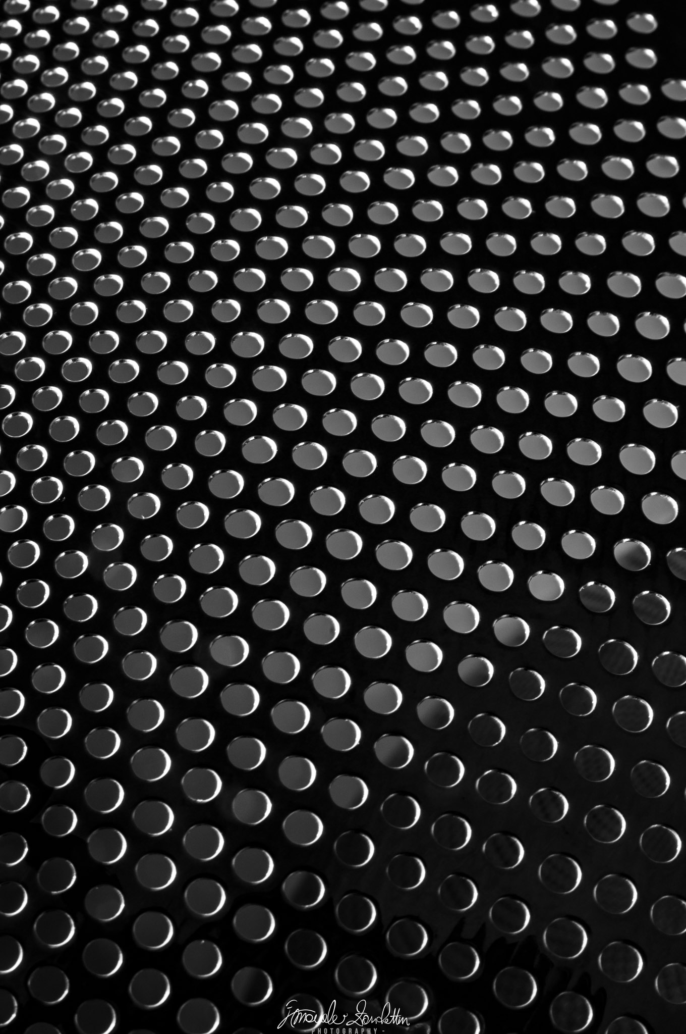 File di fori circolari riempiono una superficie scura in bianco e nero, con bordi luminosi e lievi differenze di dimensione apparente nel fotogramma.