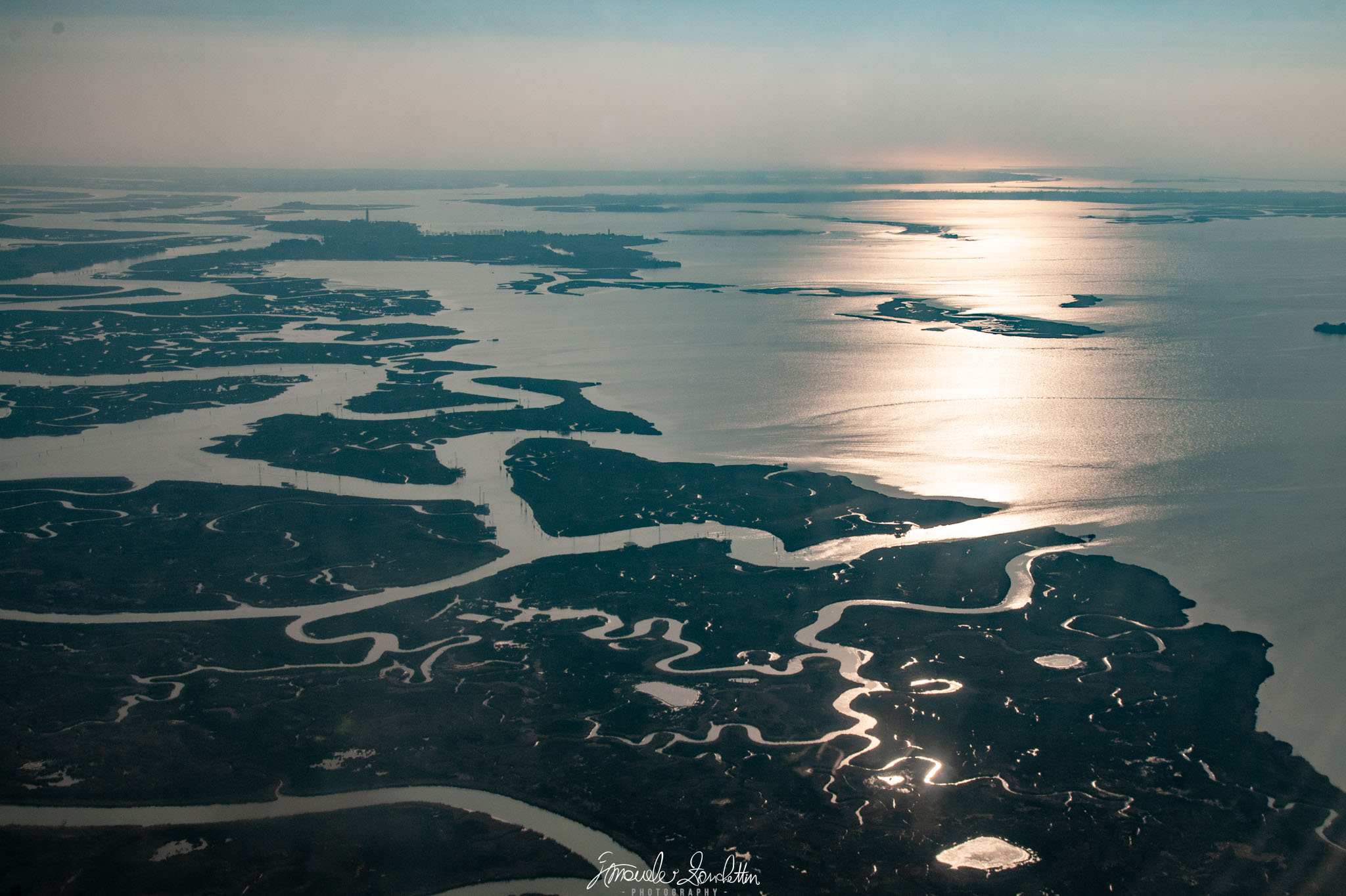 An elevated view of dark islands and branching waterways, with a broad bright reflection across the water to the right and hazy land near the horizon.