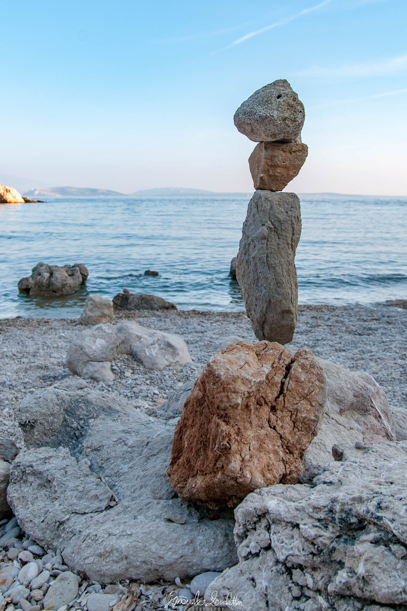 Four uneven stones form a tall stack on a rocky shore, with blue water, a distant coastline and pale sky behind them.