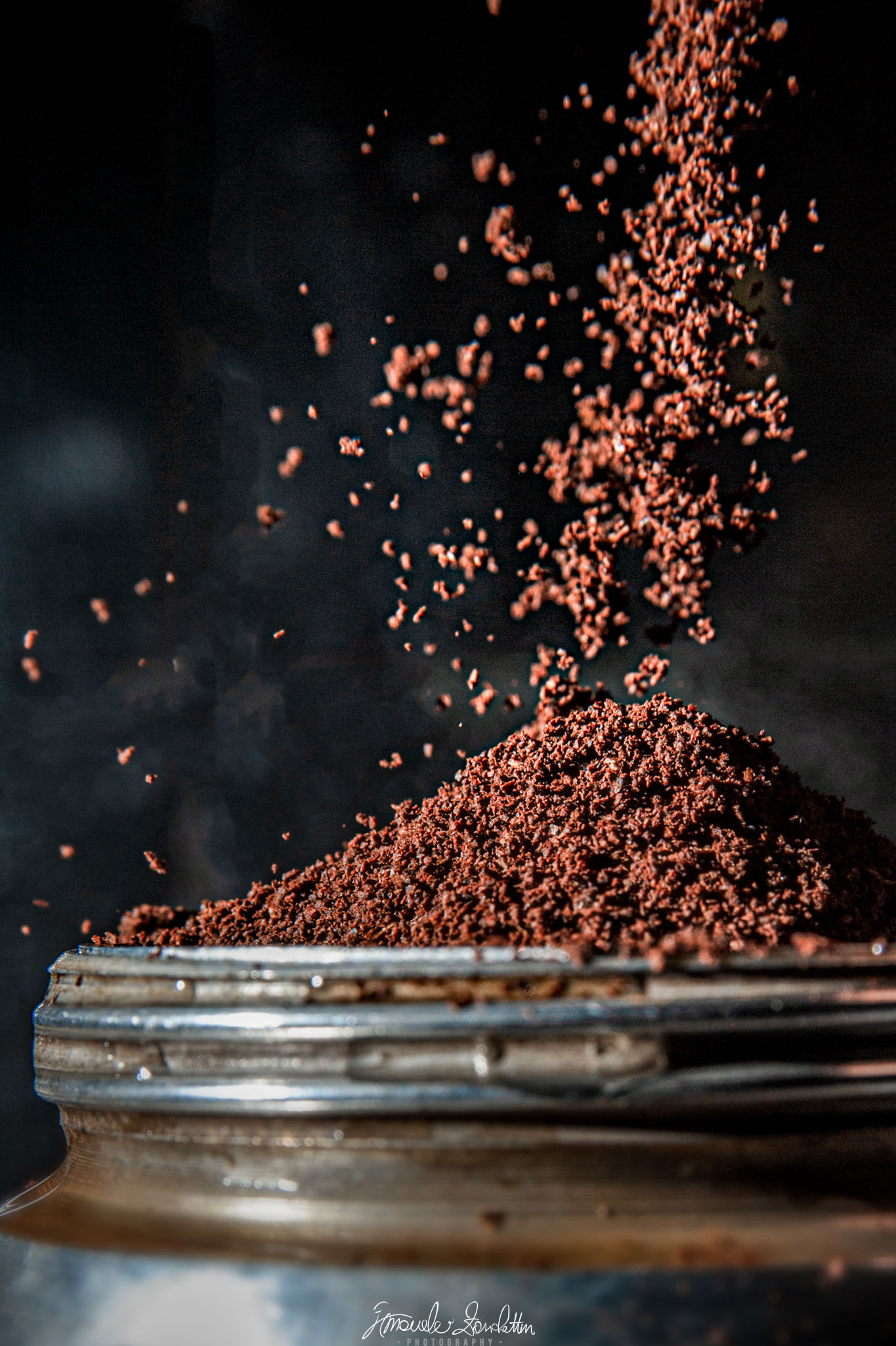 Caffè macinato marrone cade su uno sfondo scuro formando un mucchietto in un contenitore metallico, con granelli sparsi sospesi sopra il bordo.
