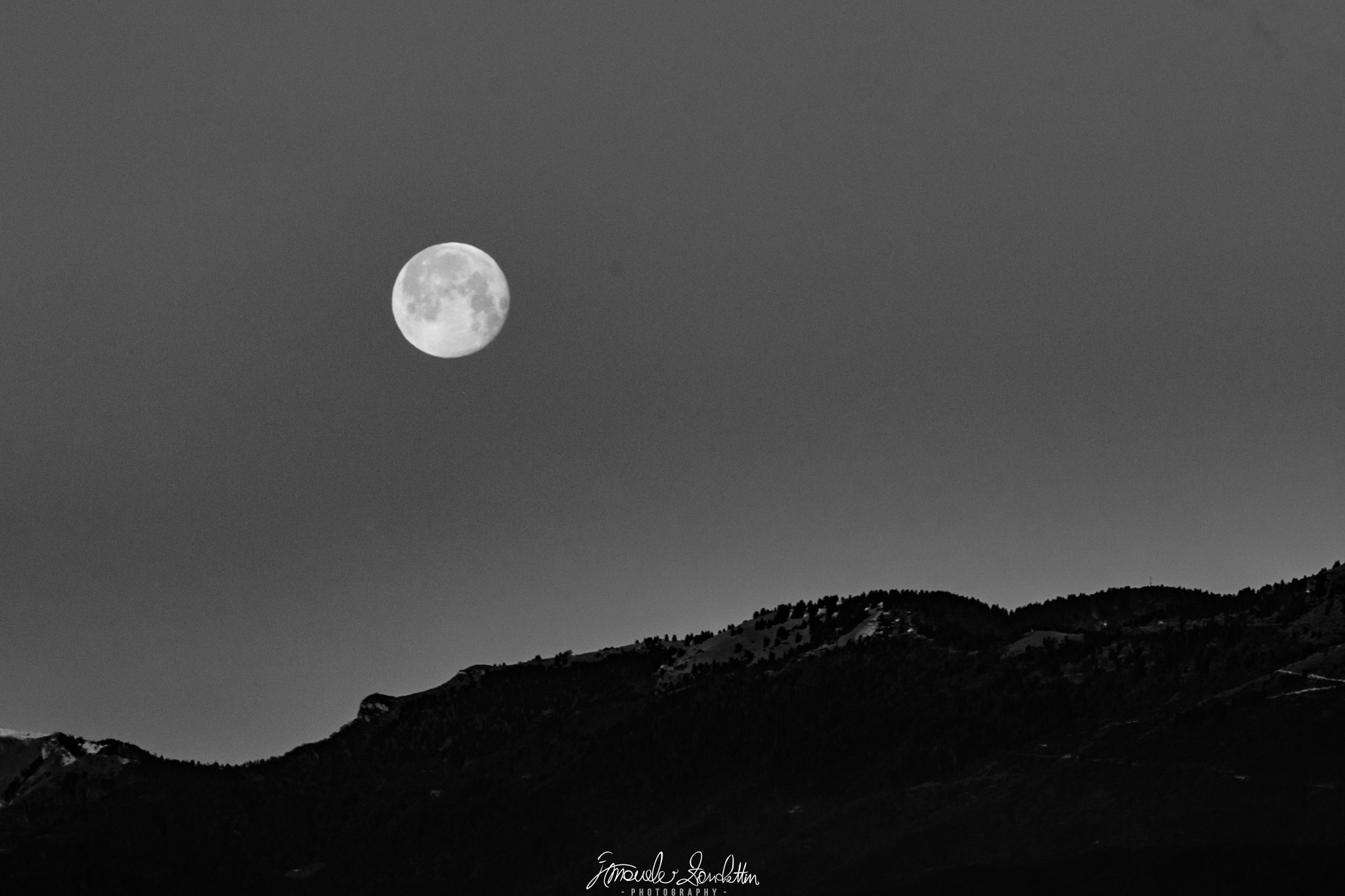 Una luna chiara si trova in un ampio cielo grigio sopra una cresta montuosa scura segnata da alberi, in una fotografia monocromatica.