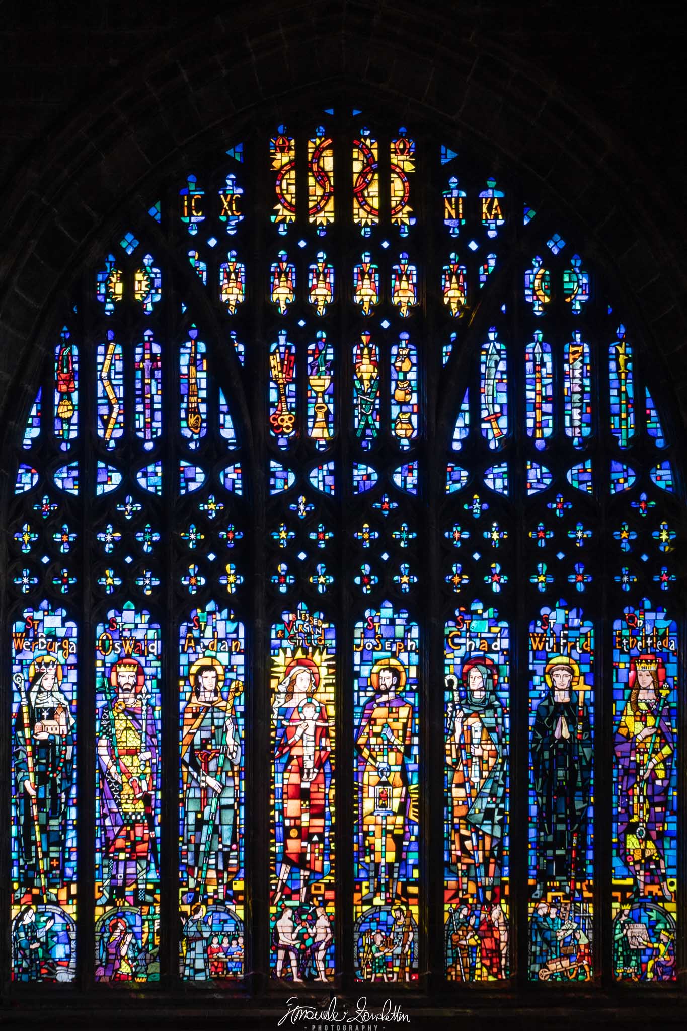 A tall arched stained-glass window contains colourful figures and repeated small patterns, separated by dark stone tracery and surrounded by a nearly black interior.