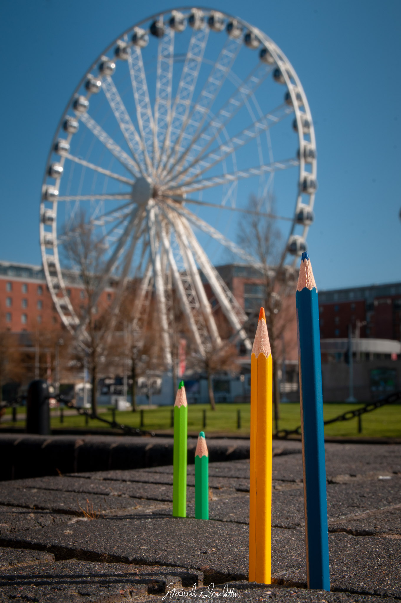Due matite alte, blu e gialla, si trovano dietro due matite verdi più piccole su una pavimentazione a Liverpool, con ombre verso sinistra e una ruota panoramica riconoscibile ma sfocata sullo sfondo.
