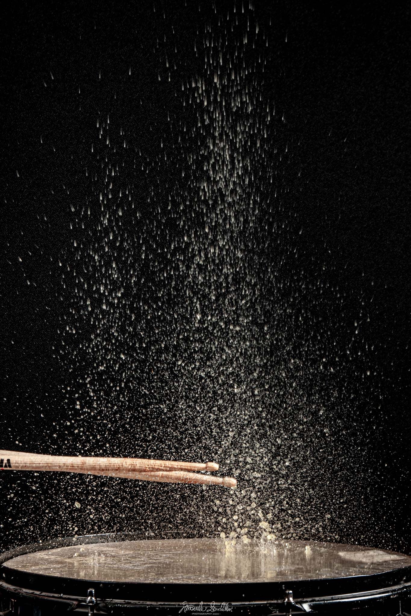 Drumsticks crossing above a wet drum skin as hundreds of water droplets are frozen against a black background