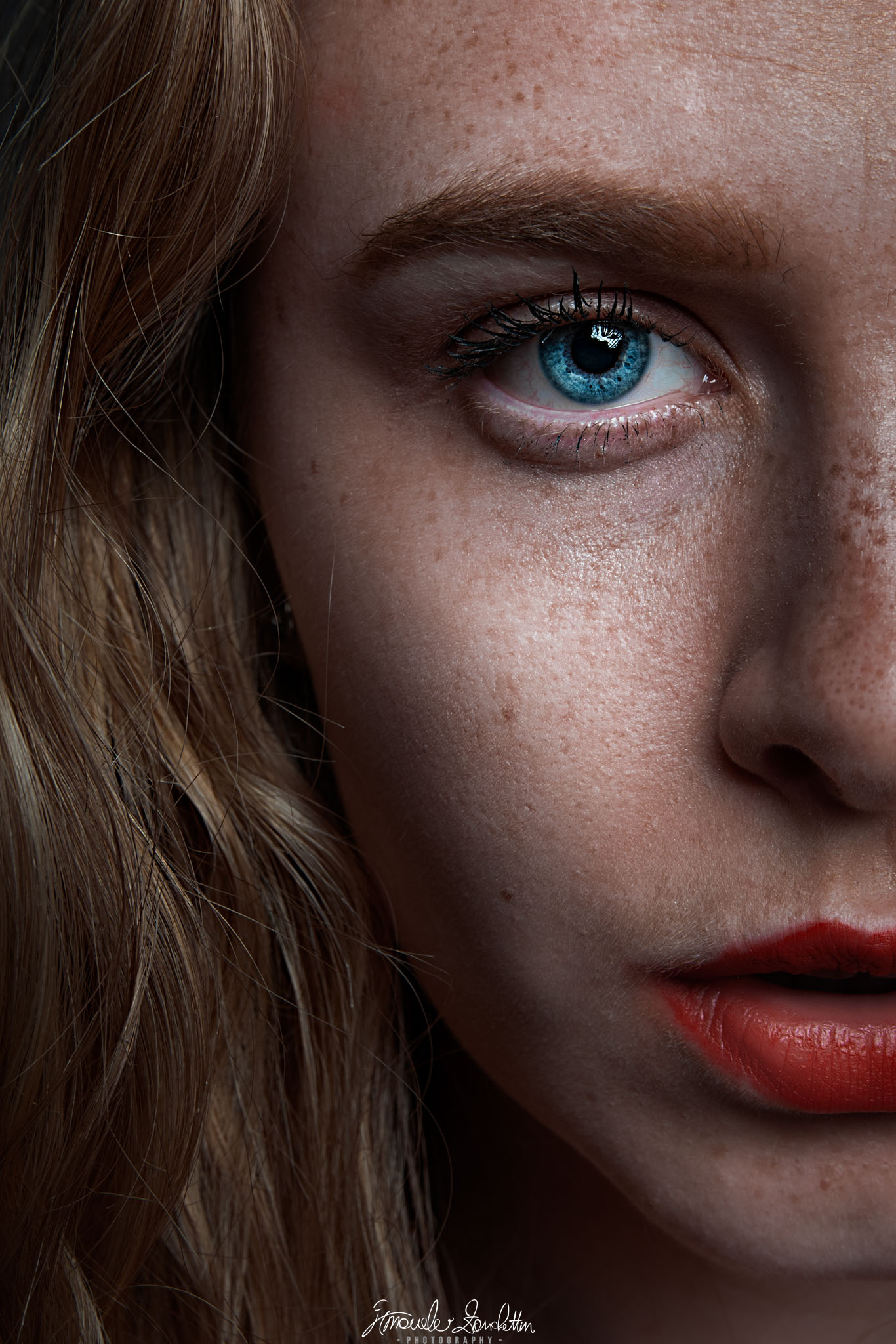Ritratto ravvicinato di una donna con capelli castani, un occhio azzurro, lentiggini visibili e rossetto rosso, con metà volto inquadrato