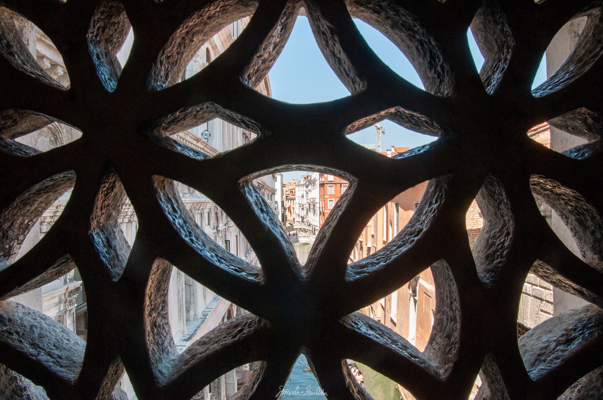 A Venetian canal and distant bridge seen through the dark repeating openings of a carved stone lattice