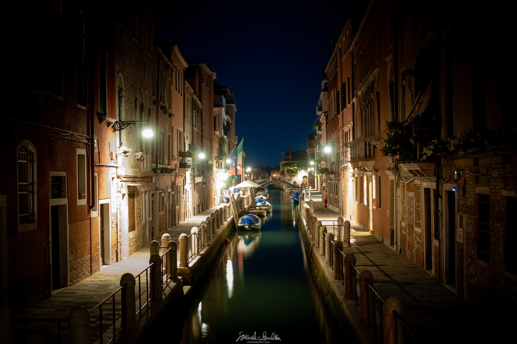 Quiet Venetian canal at night with warmly lit buildings, moored boats and reflections leading towards a small bridge