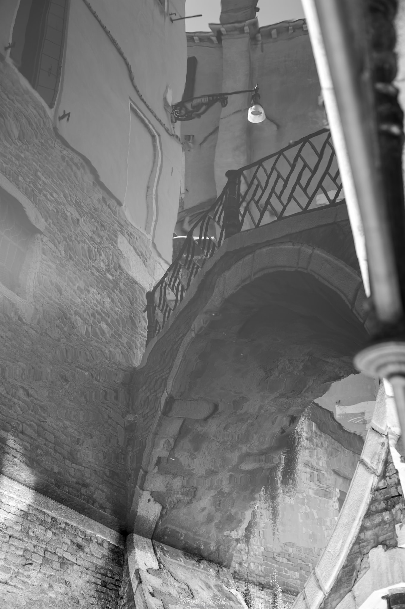 Black-and-white reflection of old walls, an arched bridge, stair rails and a hanging lamp distorted in Venetian water