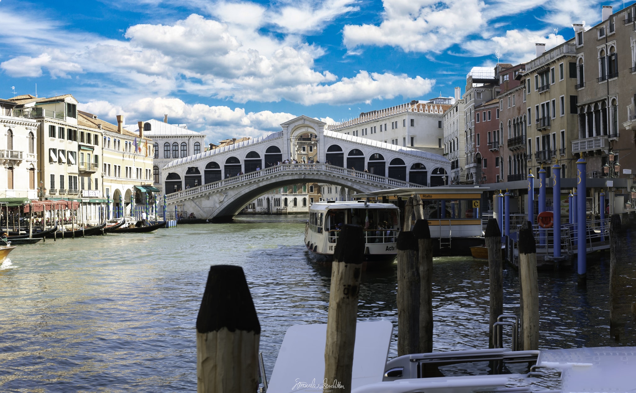 The Rialto Bridge seen across the Grand Canal with boats, gondolas, buildings and mooring posts in the foreground