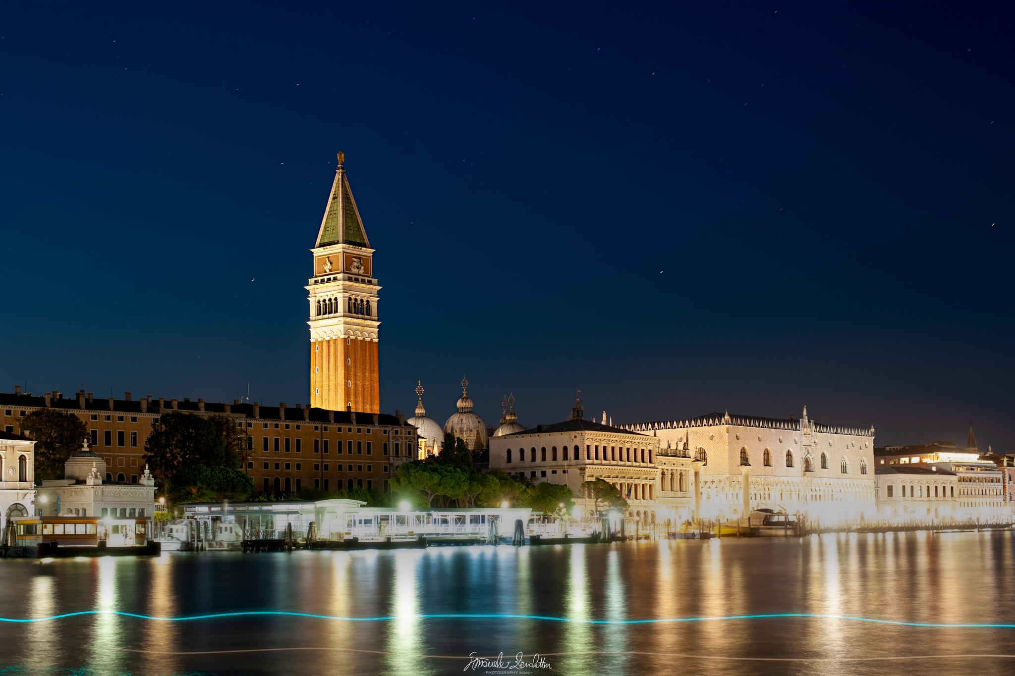 St Mark’s bell tower and waterfront illuminated at night, reflected in the lagoon with a thin blue light trail crossing the water
